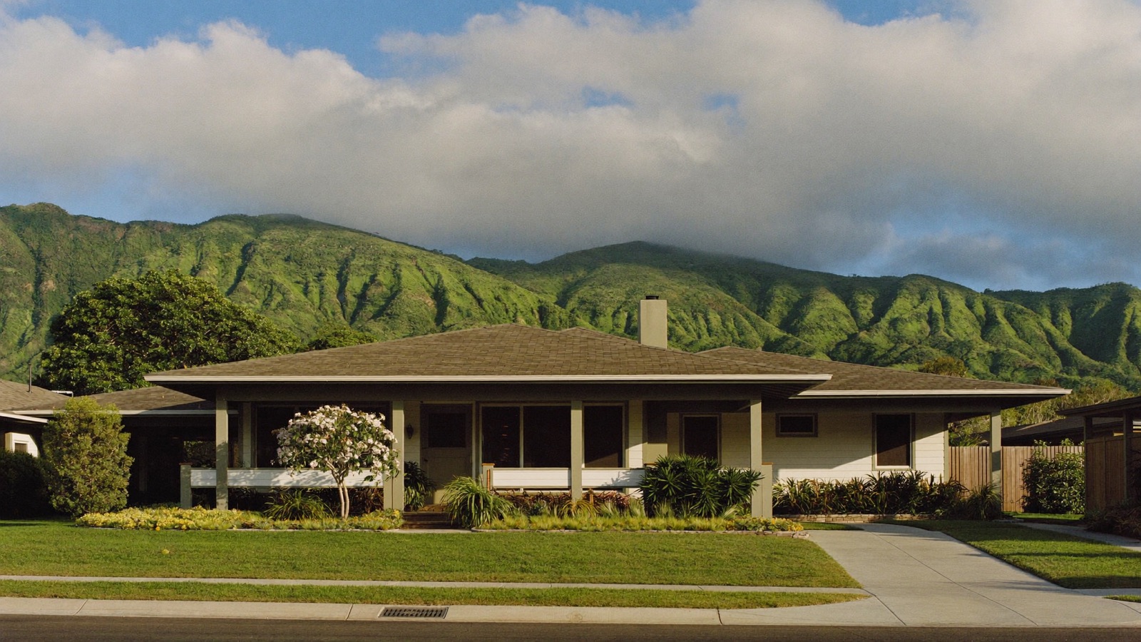 A well-kept single-story plantation-style home in an established Oahu neighborhood, with a deep lanai and a flowering plumeria, below the green Koolau ridge.