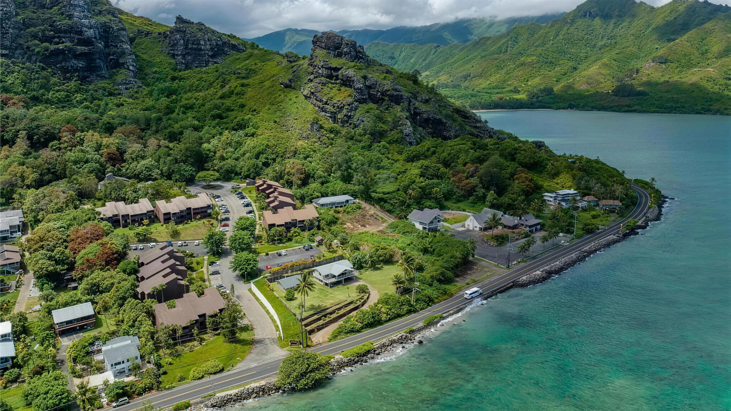 Aerial view of Makaua Village along the Windward coast: the association's brown-roofed two-storey buildings and parking areas set below a steep green ridge, with Kamehameha Highway and the turquoise shallows of the bay to the right.