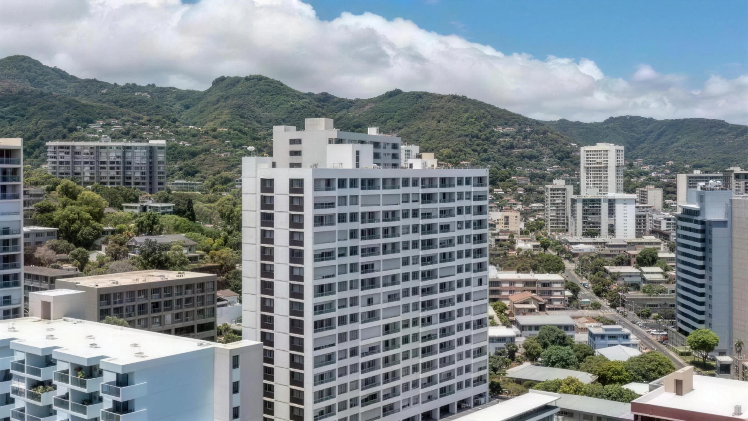 Aerial view over Punahou Royale, the white residential tower at the centre, with the low-rise Makiki neighbourhood, palm trees and the green Manoa ridgeline behind it.