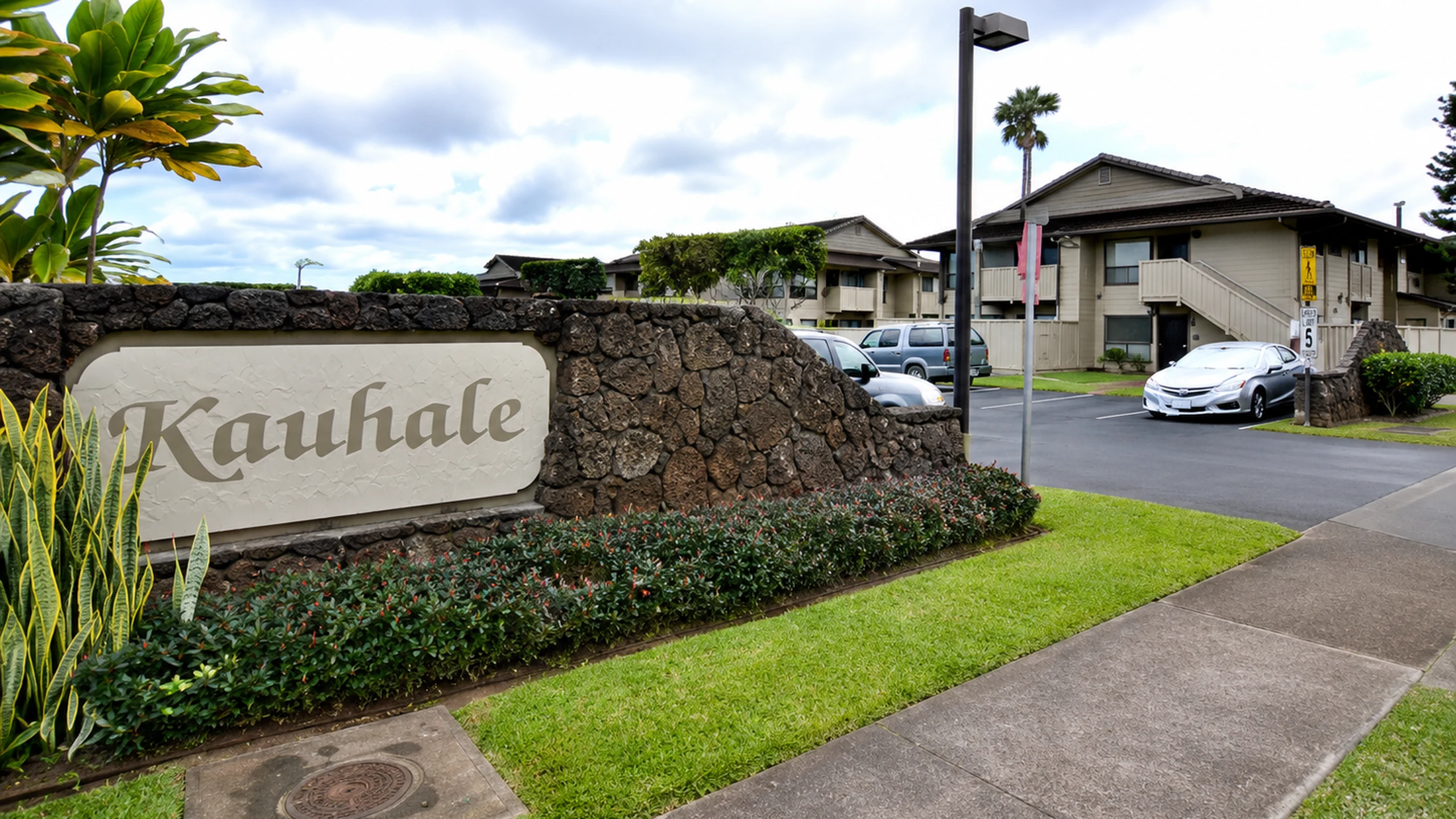 The Kauhale entrance: a lava rock wall carrying a cream sign lettered Kauhale in script, with flowering shrubs, the community's two storey townhouses and the driveway behind it.