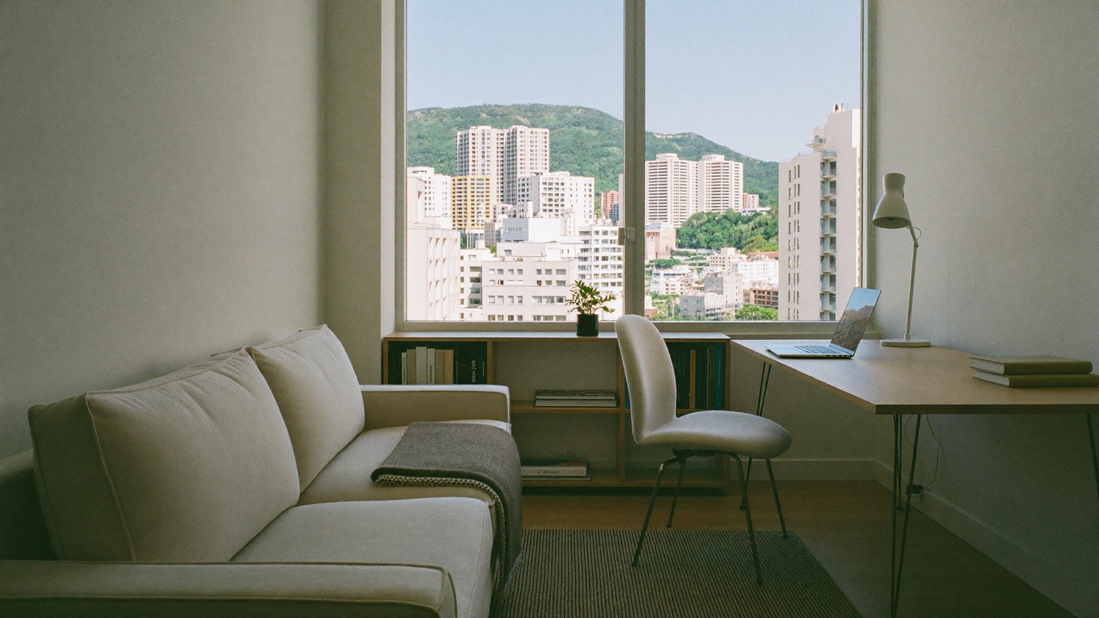 A furnished Honolulu apartment set up for a long stay, with a sofa, a work desk and laptop by the window, and urban Honolulu towers below a green ridge outside.