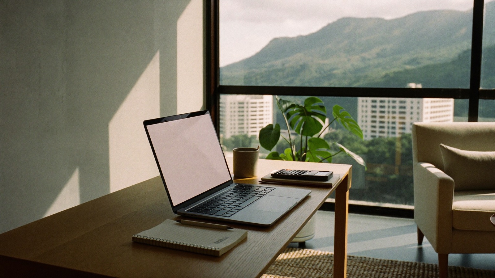A pale oak desk in a Honolulu condominium with an open laptop, a calculator, a notebook and a coffee mug, with the green Koolau ridge and Honolulu apartment towers visible through the window.