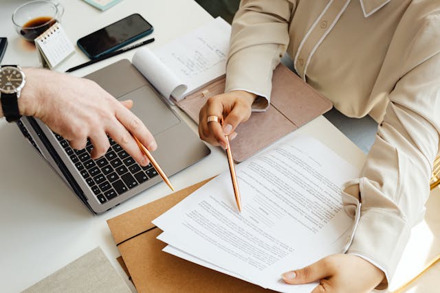 Two people reviewing paperwork at a desk with a laptop.