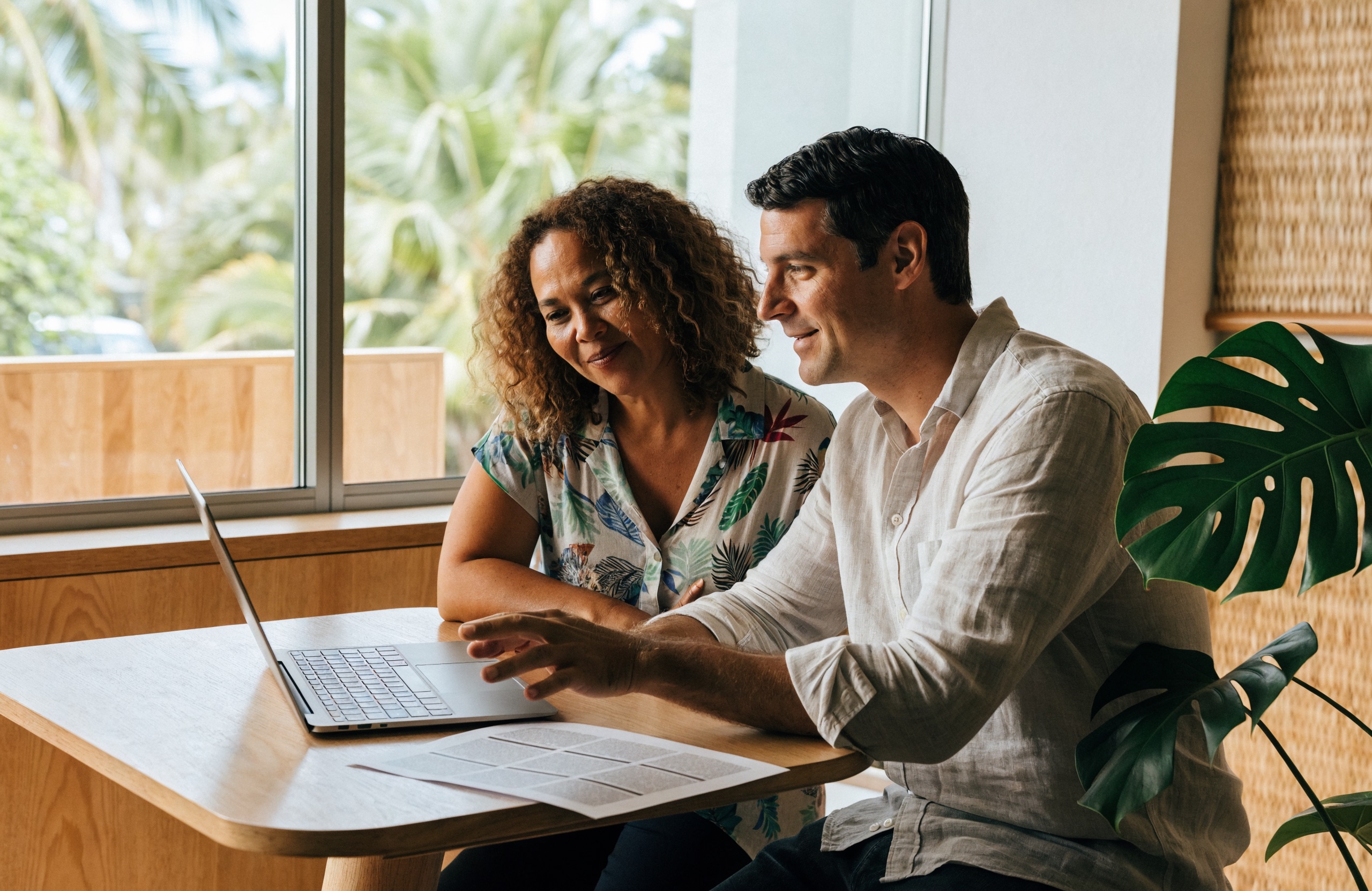 A Honolulu property manager and a rental property owner sitting together reviewing a property report on a laptop in a bright, sunlit office.