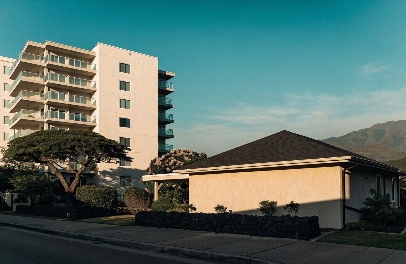 A mid-rise residential condominium with glass balconies standing directly beside a single-story Honolulu home with a hipped roof, carport and lava-rock garden wall, with a green ridge behind.