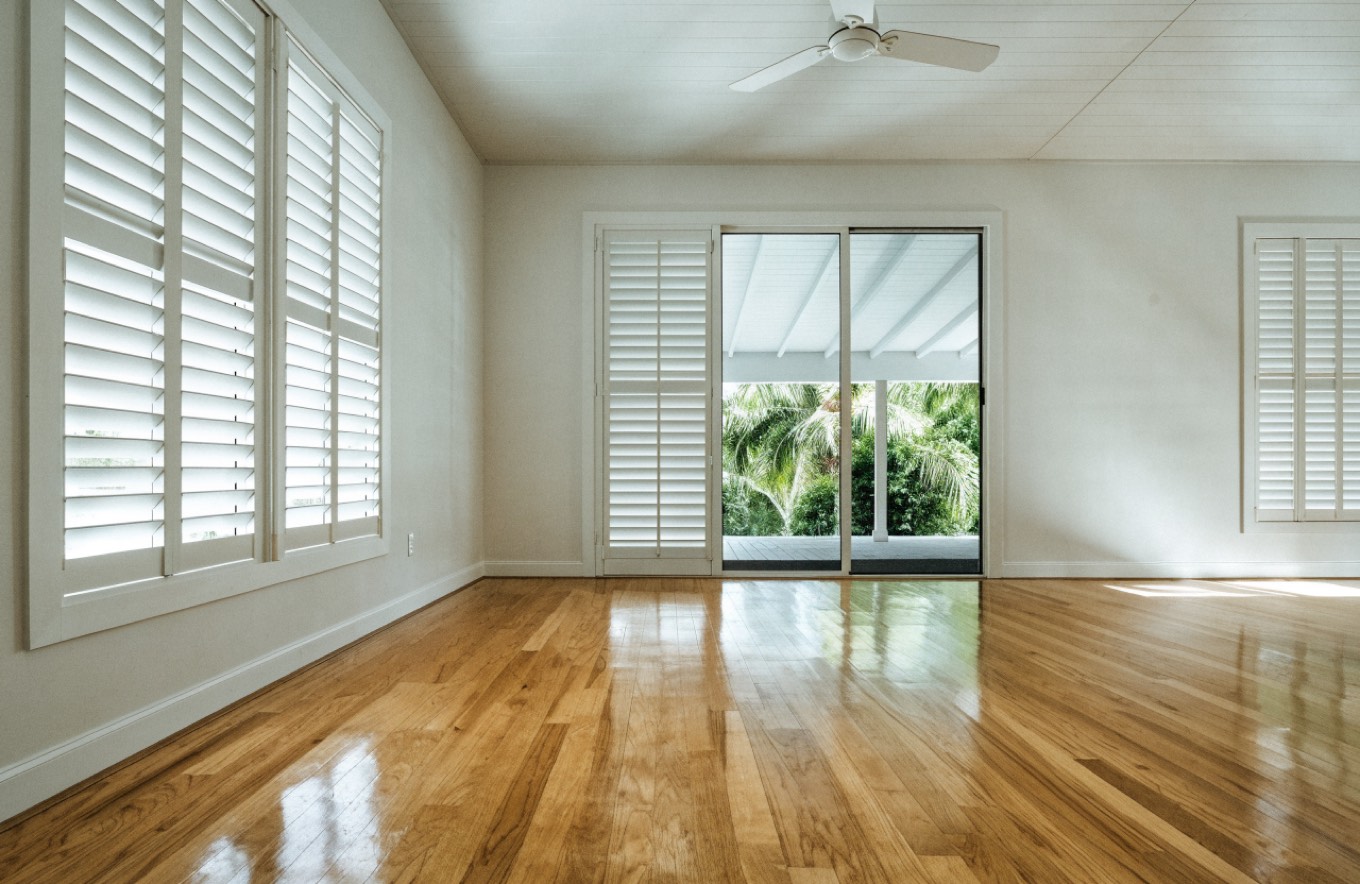 Empty living room in a Hawaii home with bare wood floors, white louvered shutters and an open door to a lanai, cleaned and prepared for a new tenant.