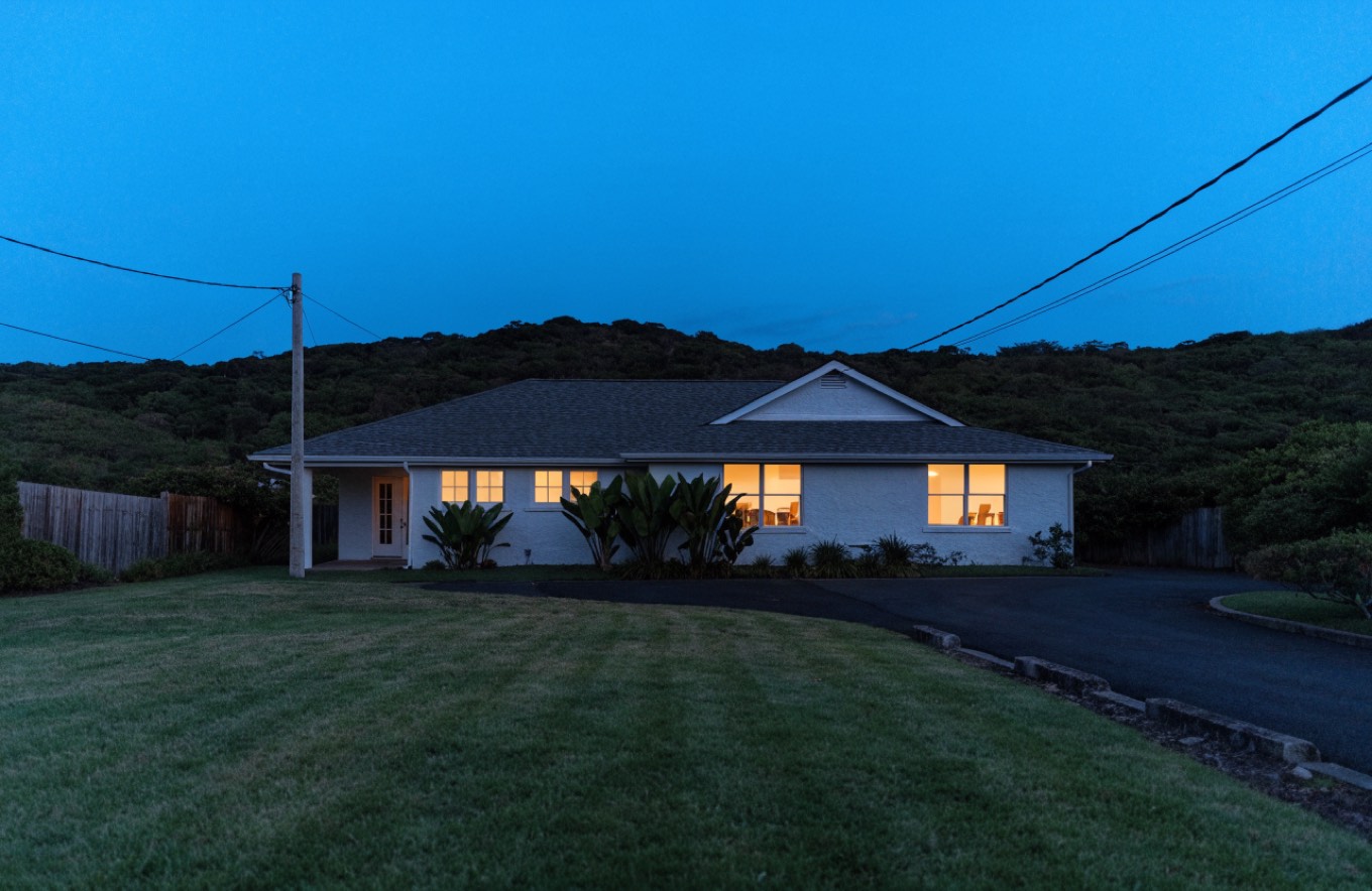 Single-story Oʻahu home at dusk with warm light showing in the windows, a wide lawn in front and a wooded ridge behind.