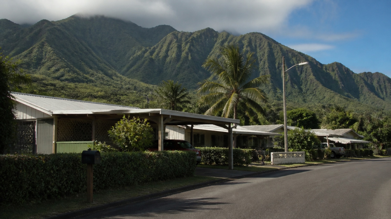 Street of single-story plantation-style homes with carports and hedges in a windward Oahu neighborhood, with the green Koolau ridgeline rising behind them.