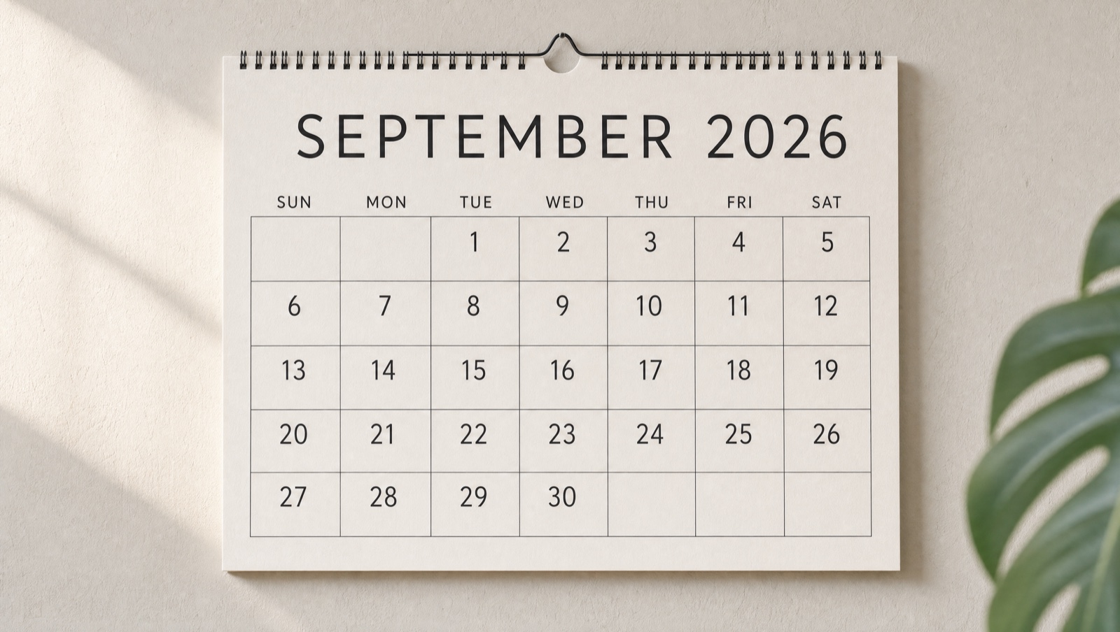 A printed September 2026 wall calendar hanging on a pale wall in soft daylight beside a monstera leaf. The month runs Sunday to Saturday with the 1st falling on a Tuesday and the 30th on a Wednesday.