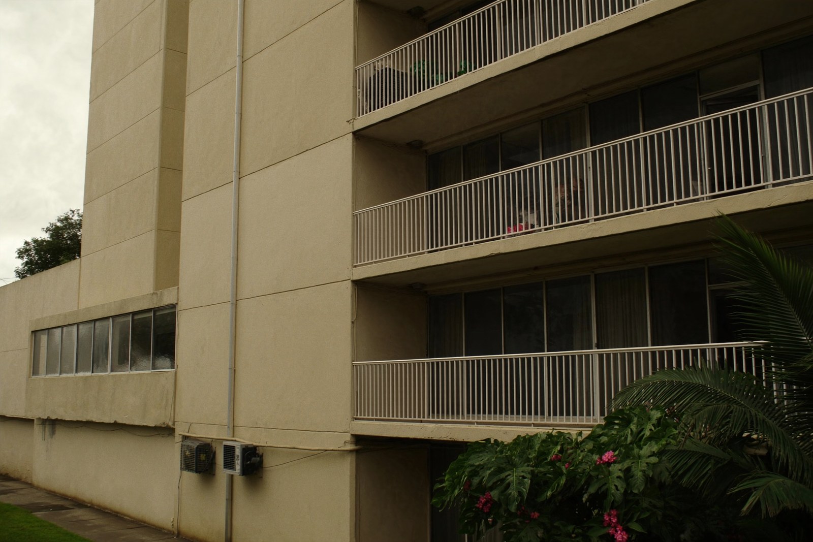 The lanai stacks of a Honolulu residential condominium tower in painted concrete under an overcast sky, with mature tropical planting at the base.