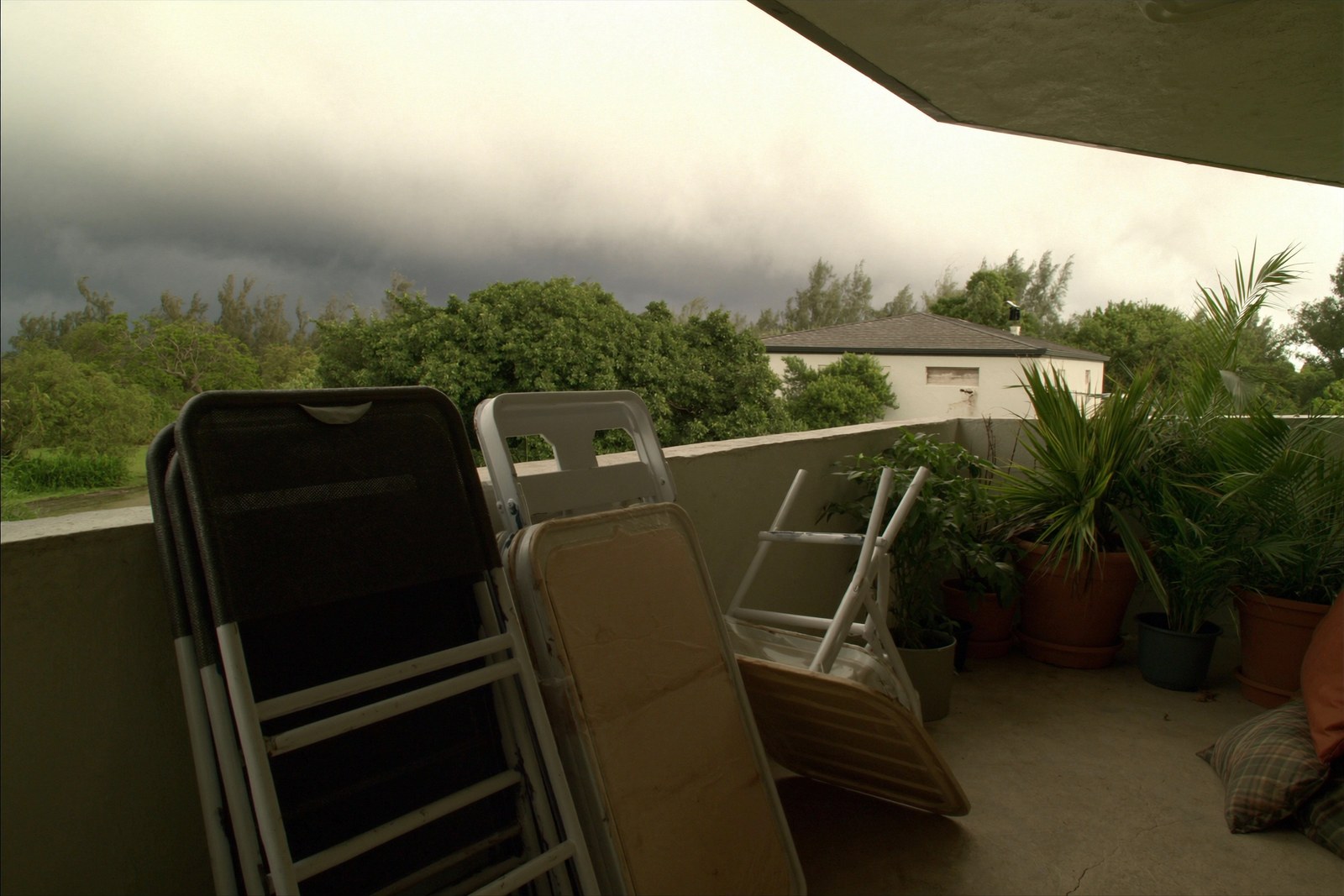 Folded outdoor chairs stacked against the wall of a residential lanai and potted plants pulled in close to the building, with dark storm cloud building in the distance.