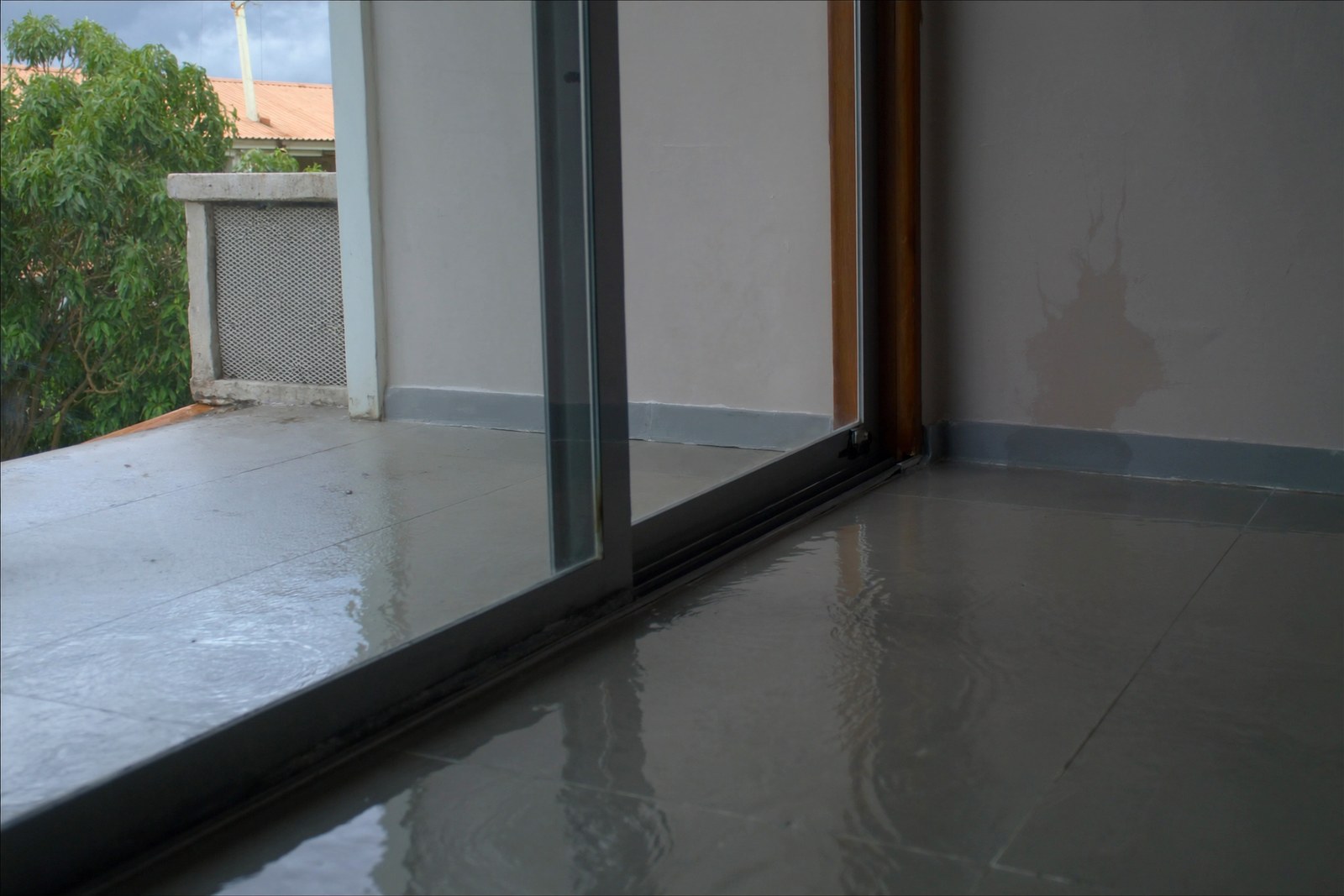 Rainwater pooled on a tiled lanai and tracking under a sliding glass door onto the floor inside, with a damp patch spreading up the base of the interior wall.