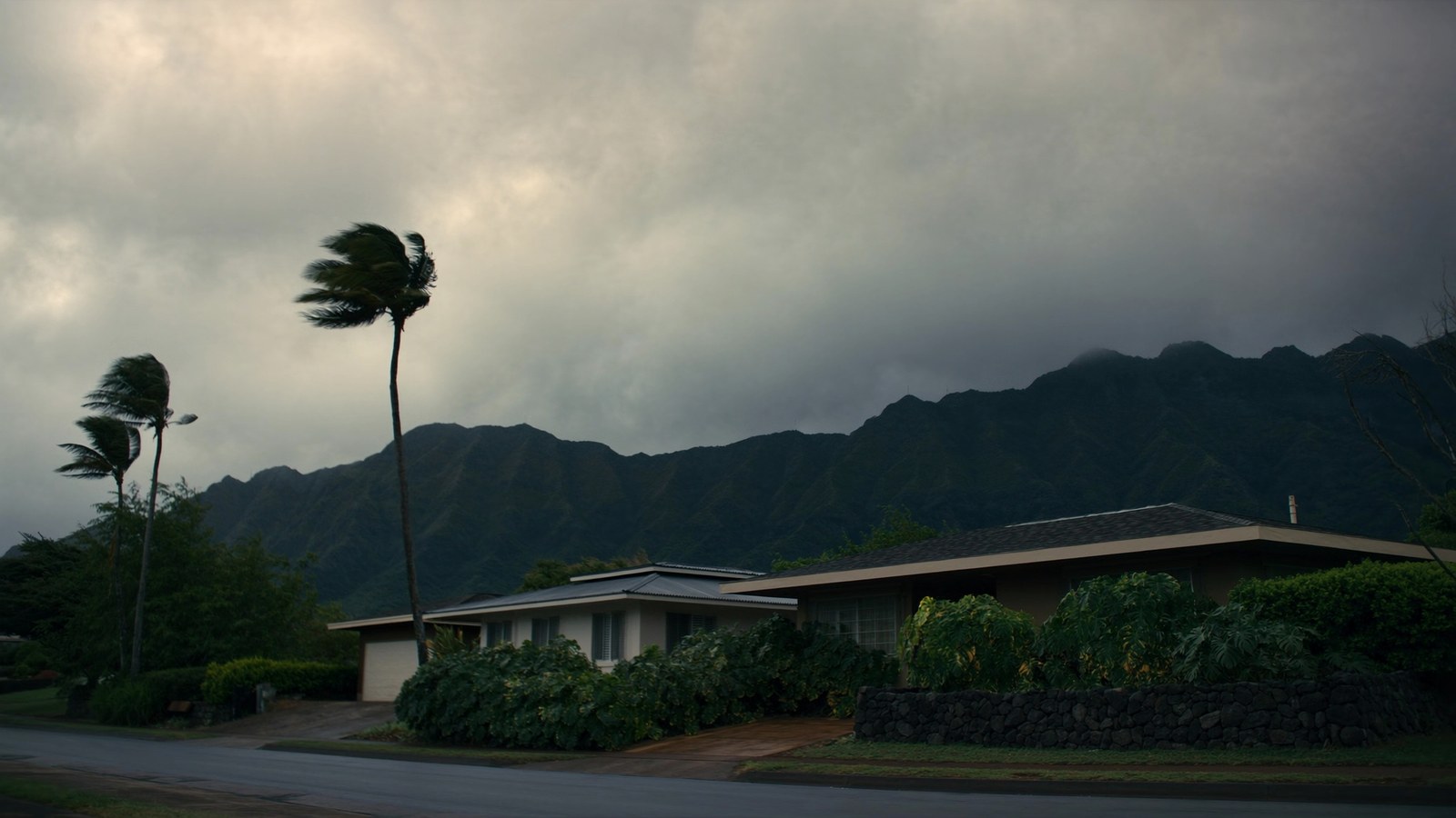 A street of low-rise Oʻahu homes with carports and hedges under a heavy storm cloud building over the green ridgeline behind, the pavement already wet from rain.