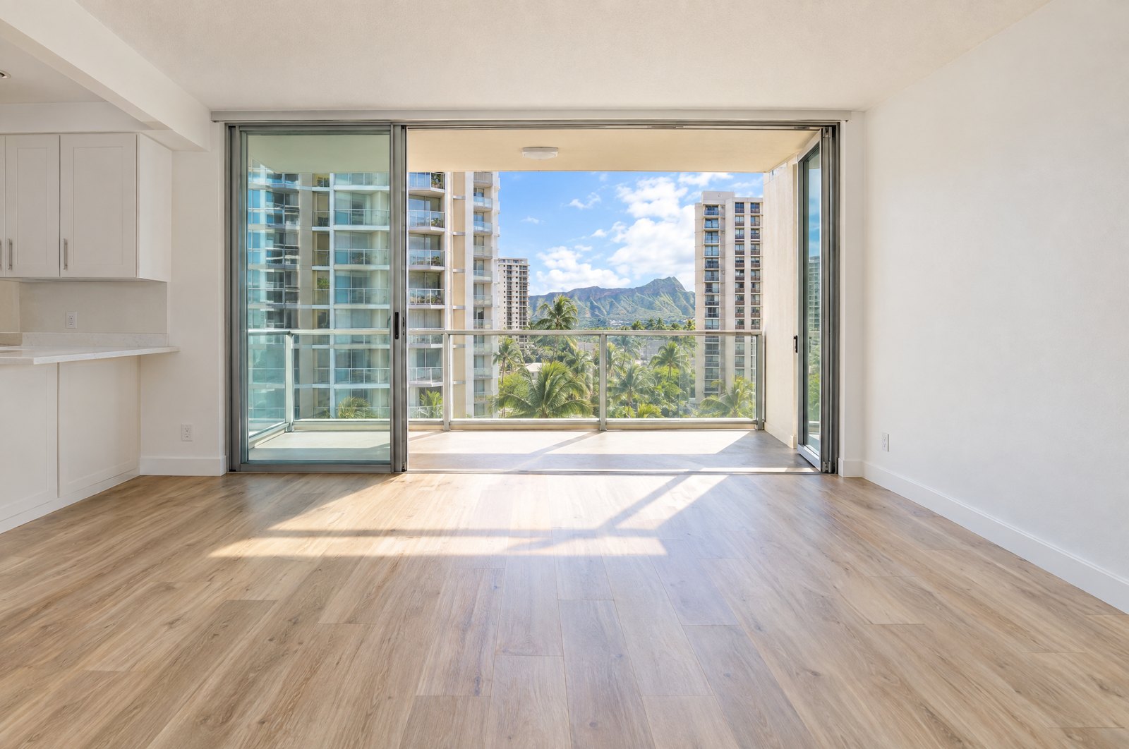An empty, freshly cleaned condominium living room with wood-look flooring, its sliding glass door open onto a lanai that looks out at neighboring towers, palm tops and Diamond Head.