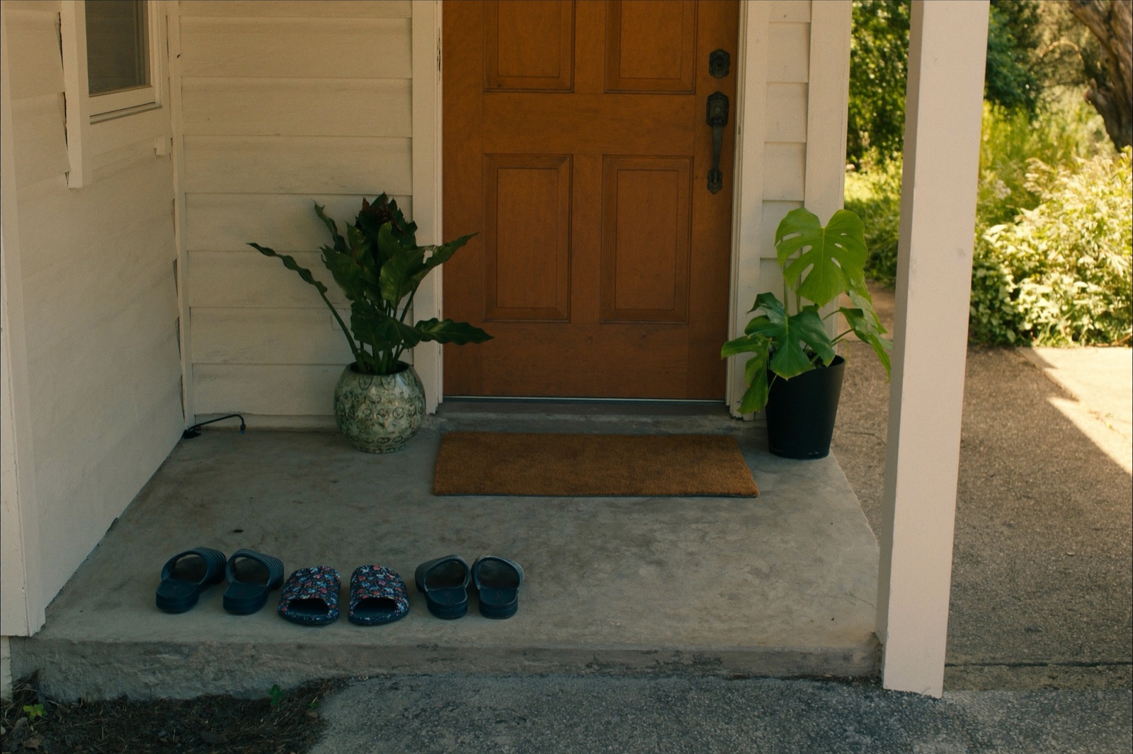 The front step of a single-family home with a wooden door, a plain doormat, potted plants on either side, and three pairs of slippers lined up on the concrete.