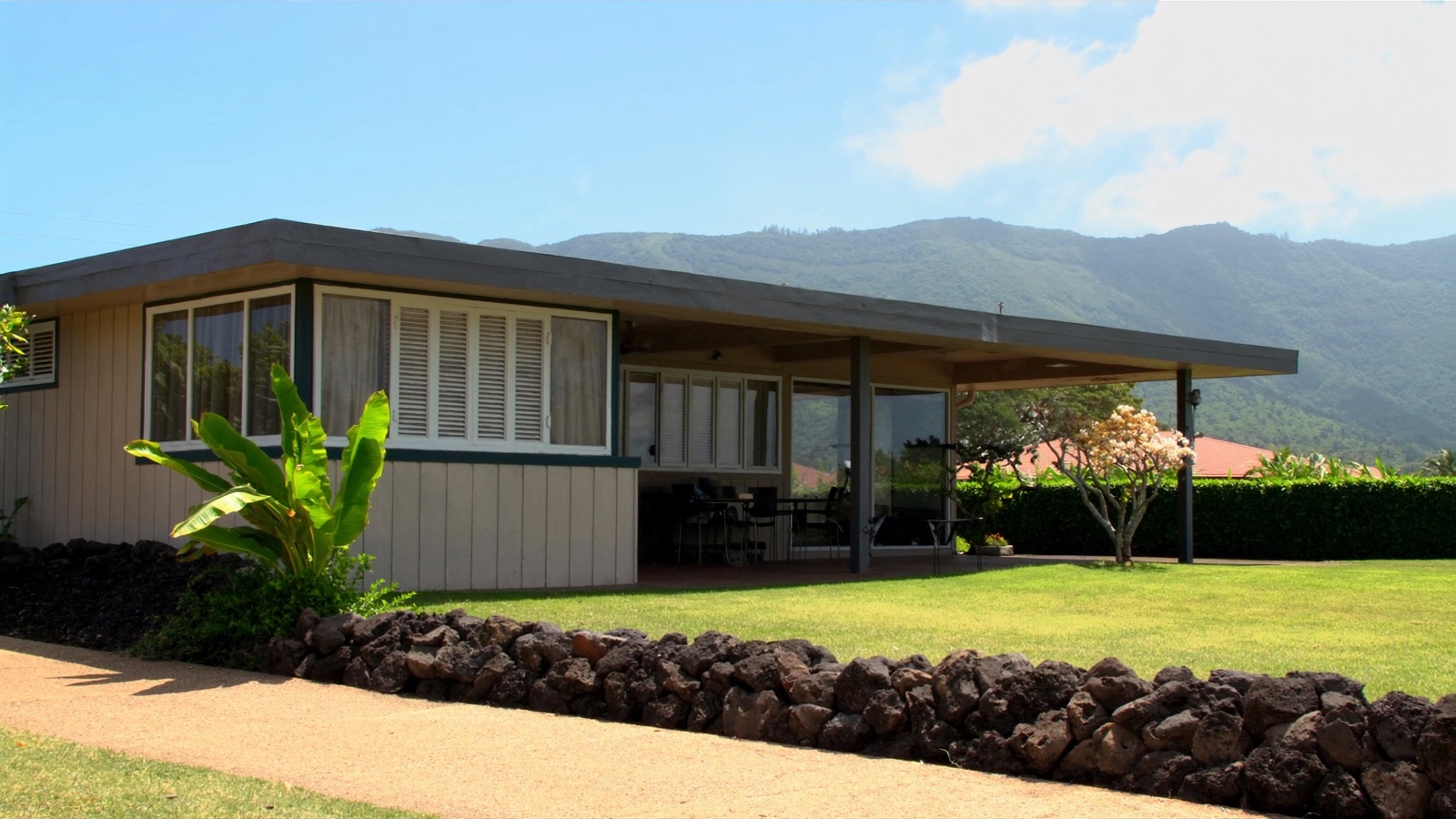 A single-story Oʻahu house with pale wood siding, white jalousie windows and a covered lanai, a low lava rock wall and green lawn in front, and the green Koʻolau ridge under a bright blue sky behind.