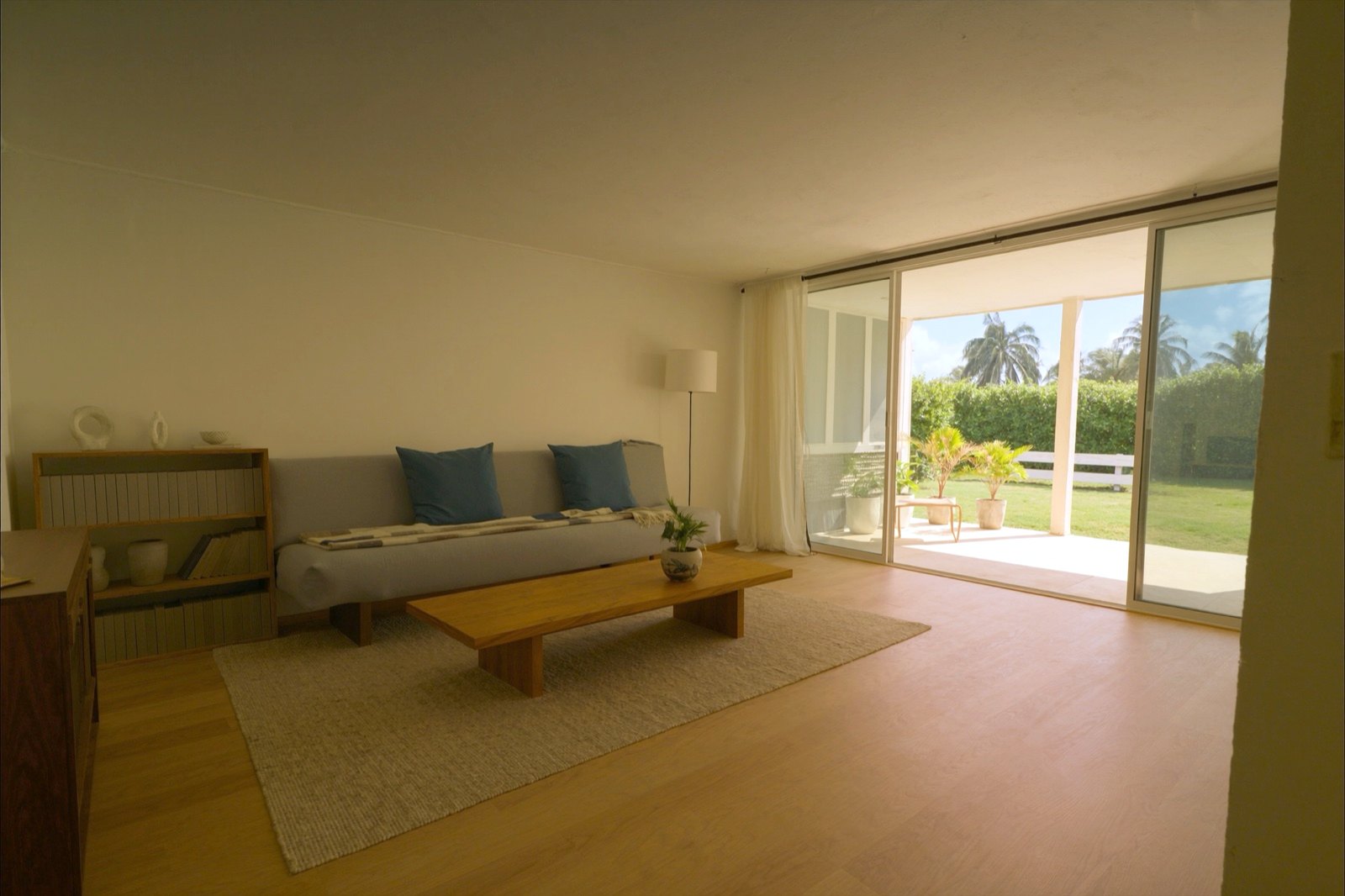 A tidy living room with a gray sofa, a low wooden coffee table, a woven rug, a bookshelf and a floor lamp, its sliding glass door open onto a covered lanai with potted plants and a green yard beyond.