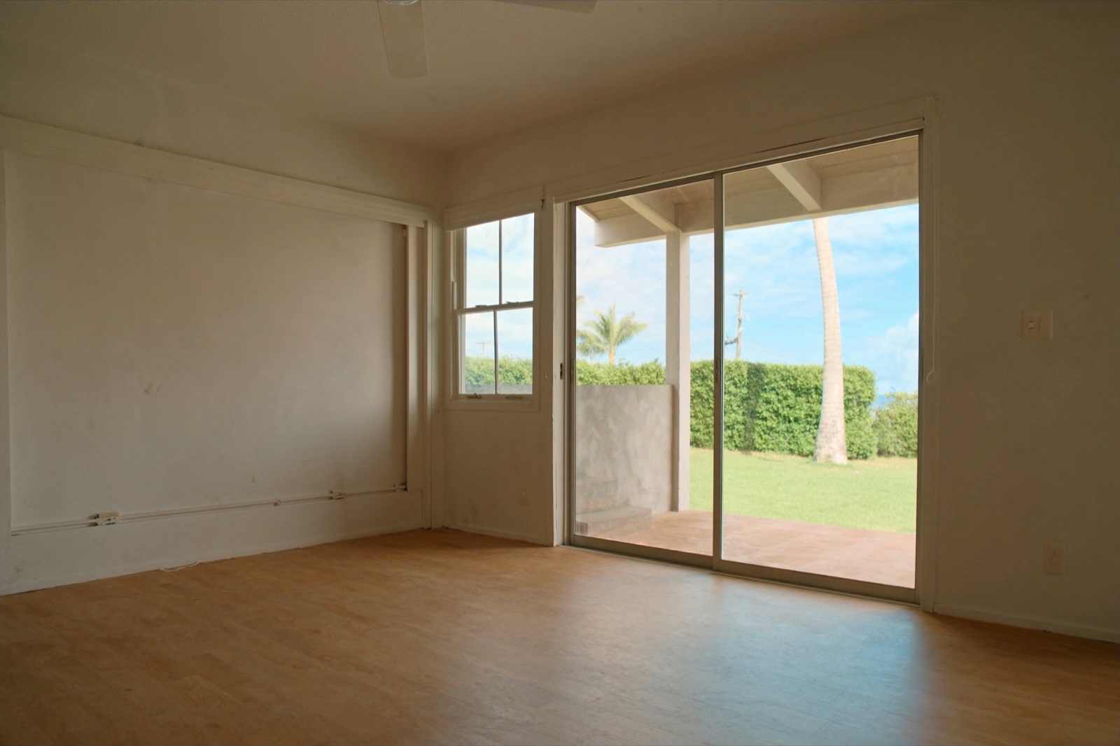 An empty living room after move-out, with a bare wood-look floor, white walls and a sliding glass door open to a covered lanai, a lawn, a hedge and a palm tree.