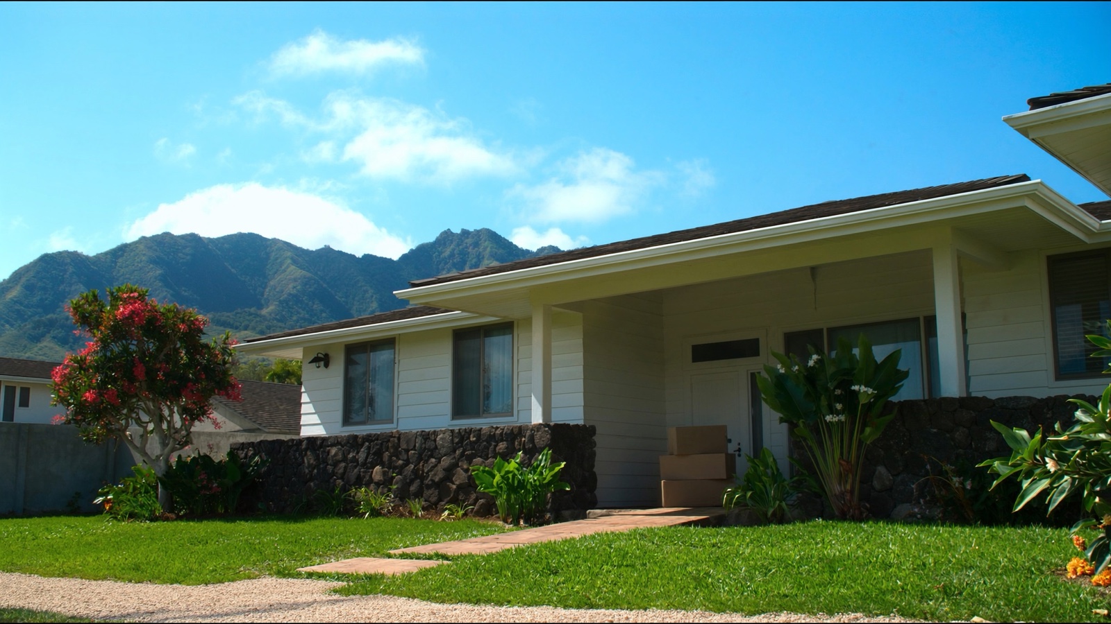 A single-story Oʻahu house with white wood siding and a covered front porch, three plain cardboard moving boxes stacked by the front door, a lava rock wall, a flowering plumeria and the green Koʻolau ridge under a blue sky.