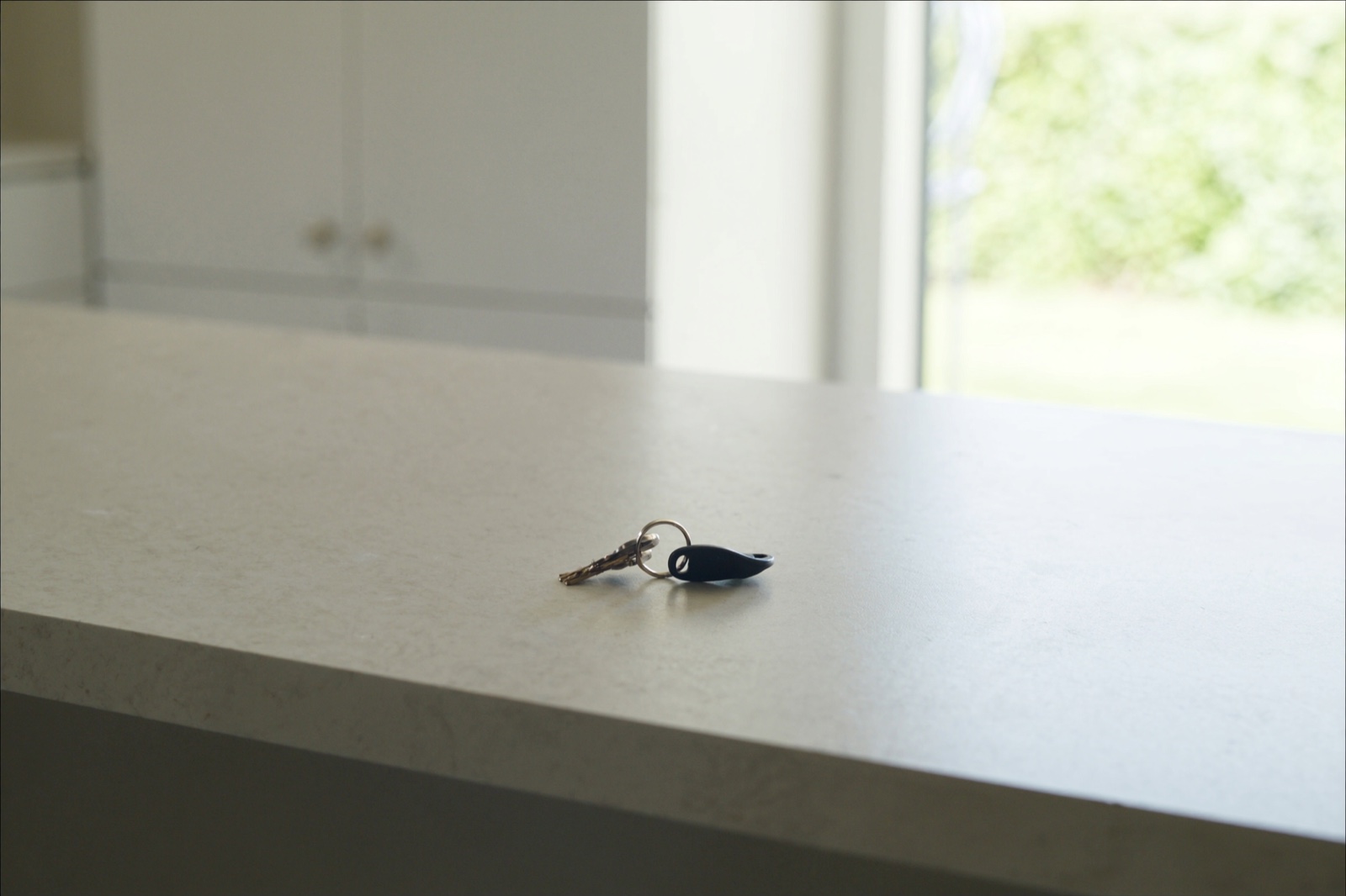 A small ring of house keys with a plain black key fob left on an empty quartz kitchen counter, with a window and green foliage behind.