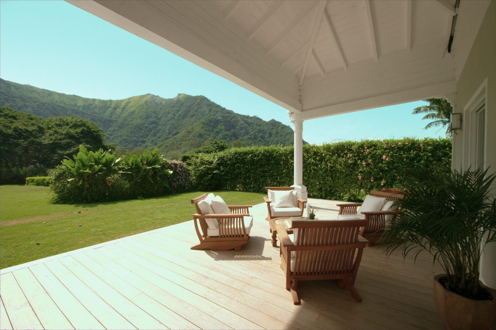 A sunny covered lanai with a white ceiling and a light wood deck, teak chairs with white cushions, a green lawn and a flowering hedge, and a green mountain ridge under a blue sky.