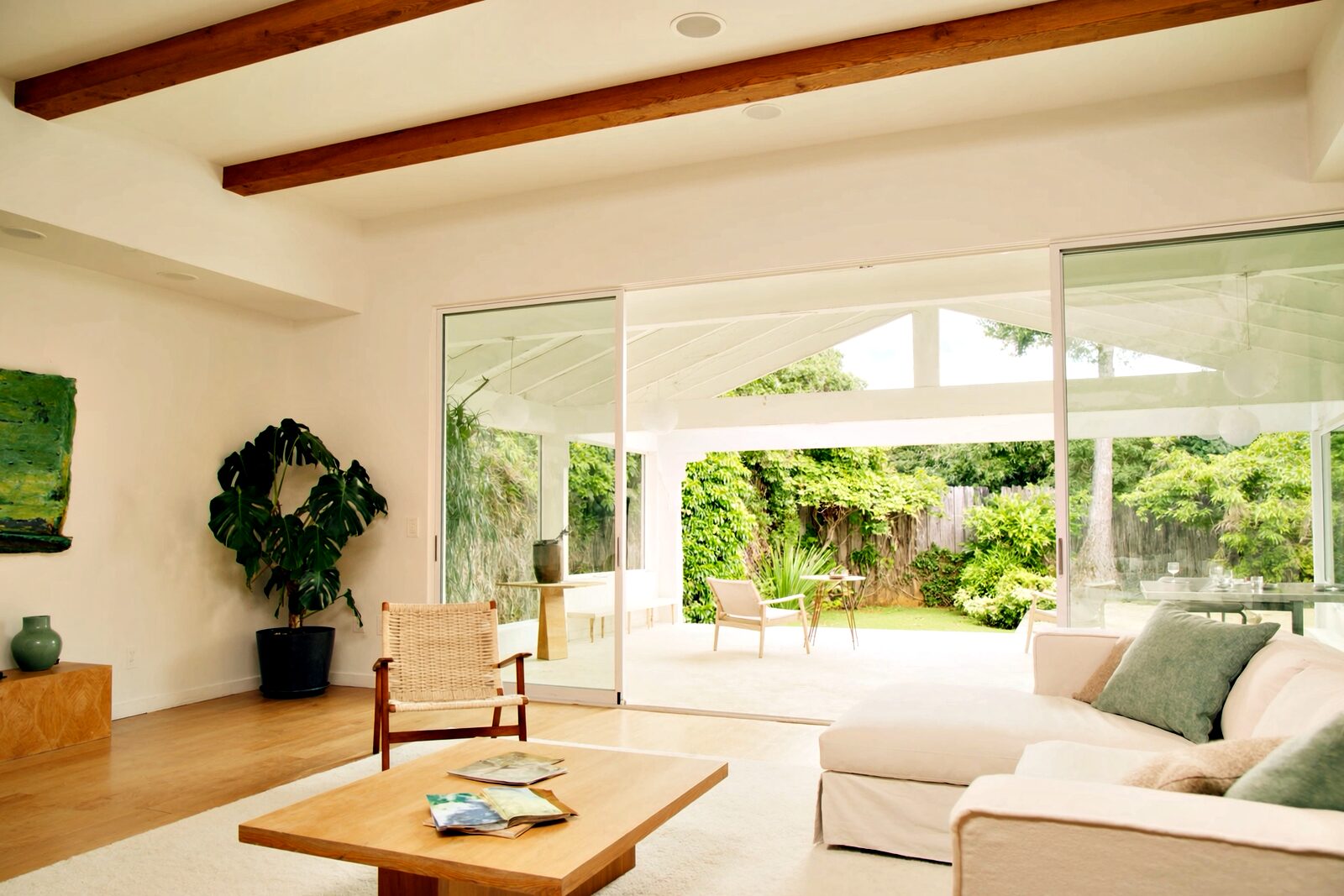A bright, airy living room with white walls, wood ceiling beams and a light oak floor, a cream sofa with sage cushions, a rattan chair and a potted monstera, opening through wide glass doors onto a covered lanai and a green garden.