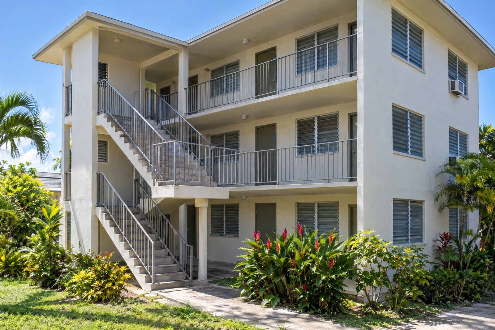 A three-story walk-up apartment building in sunshine, with open exterior concrete stairs and walkways, metal railings, jalousie windows, a window air conditioner and red ginger at its base.
