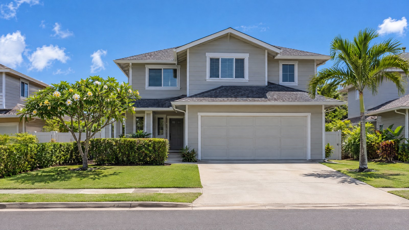 A two-story single-family home with light gray siding, a closed two-car garage and a wide concrete driveway, with a flowering plumeria, a trimmed hedge and a palm, on a sunny suburban street.